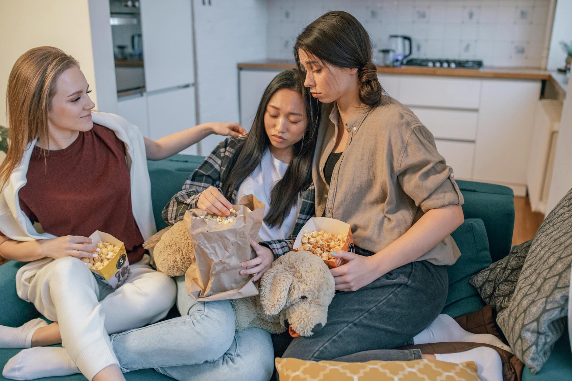 Three women share popcorn on a couch, bonding and offering support. An intimate, cozy moment indoors.