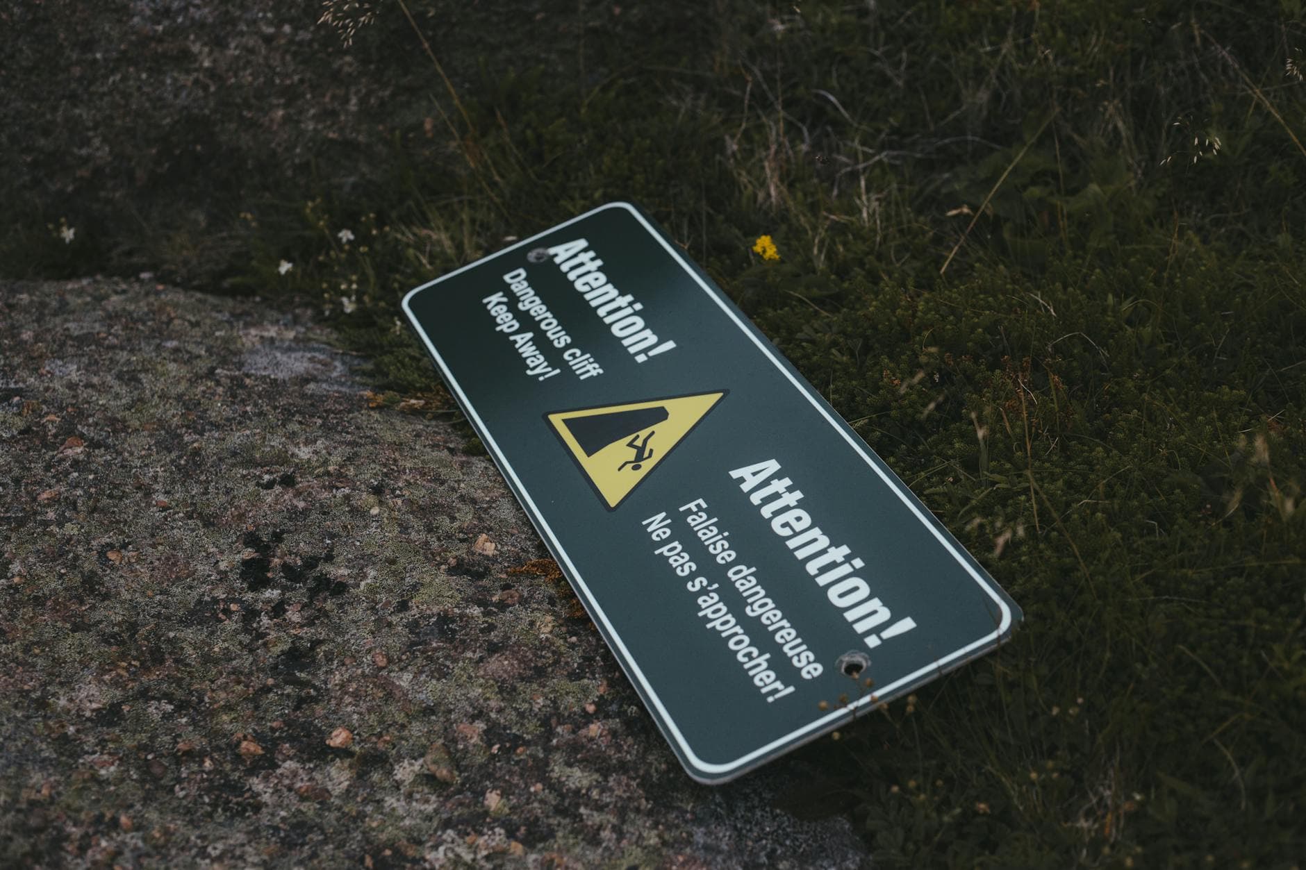An outdoor warning sign saying 'Attention! Dangerous cliff. Keep away.' lying on rocky ground.