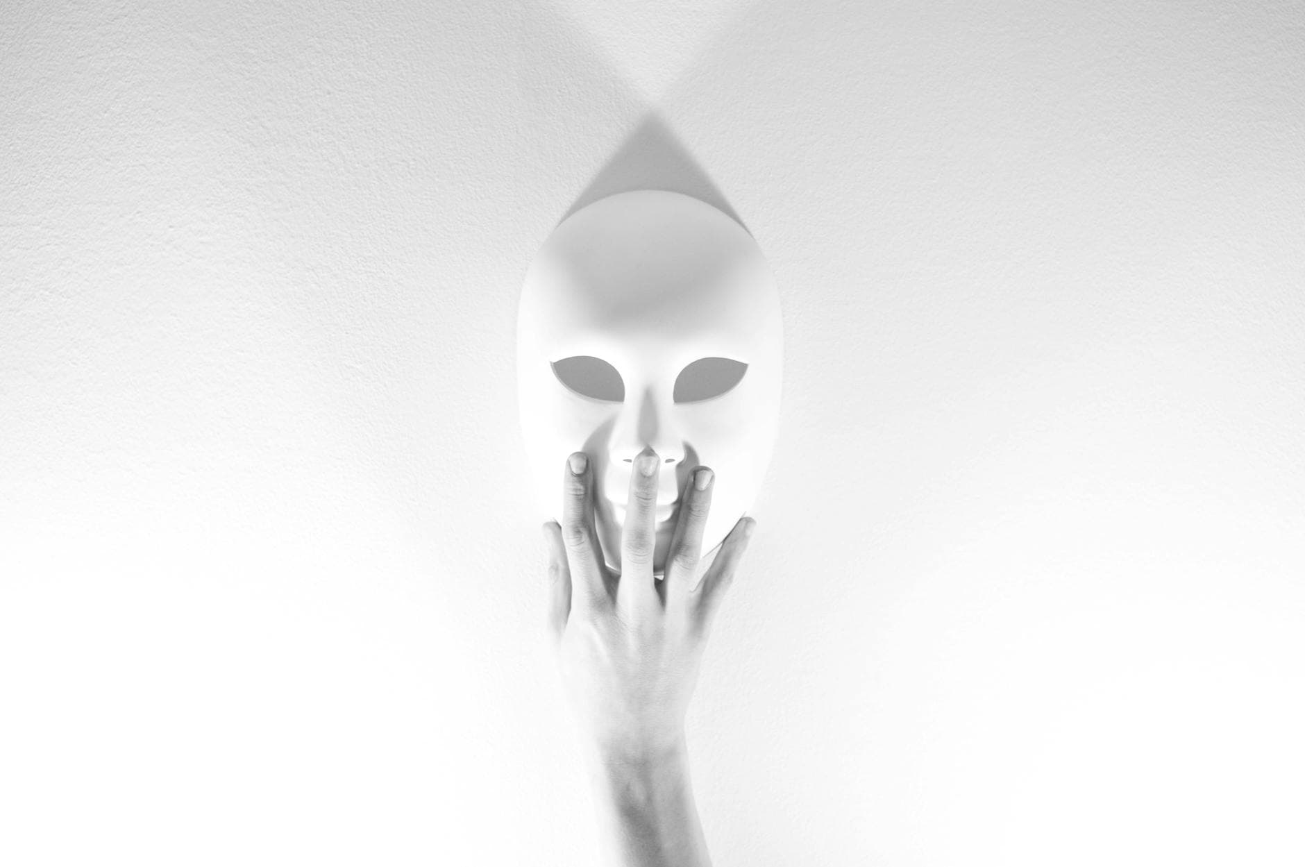 A white mask touched by a hand on a white wall, evoking mystery and introspection in a minimalist setting.