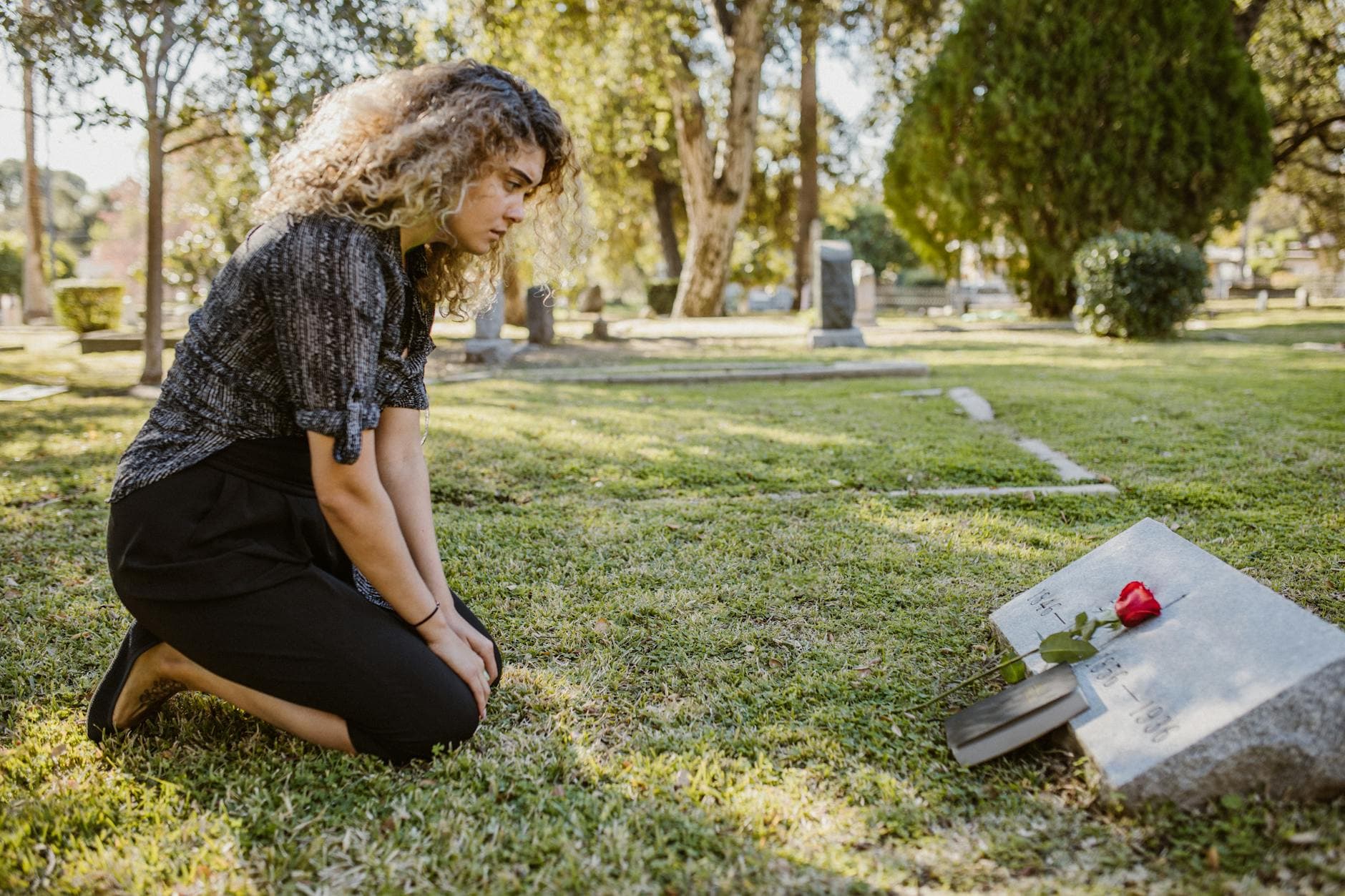 A woman kneeling by a grave in a sunlit cemetery, expressing grief and remembrance.