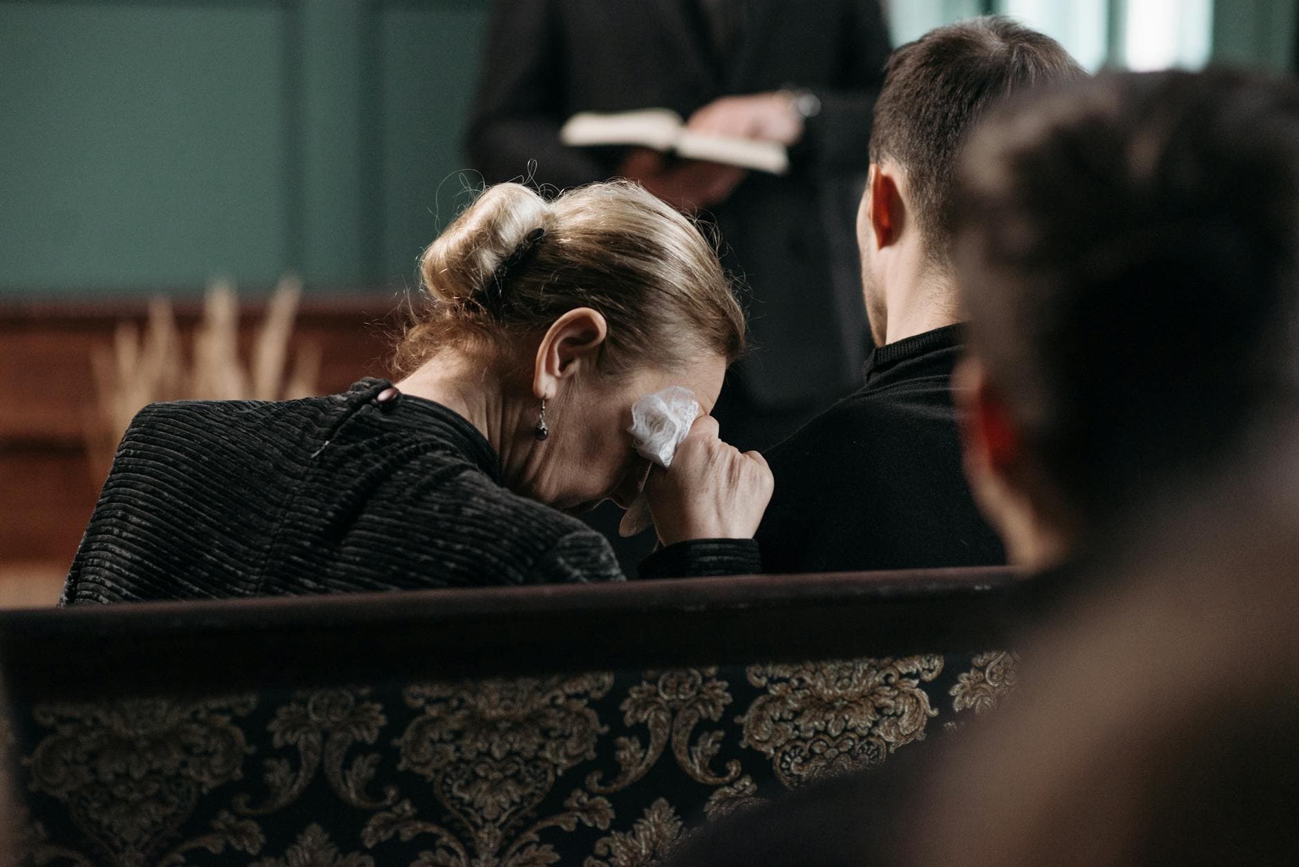 A grieving woman wipes tears during a heartfelt funeral ceremony in a somber indoor setting.