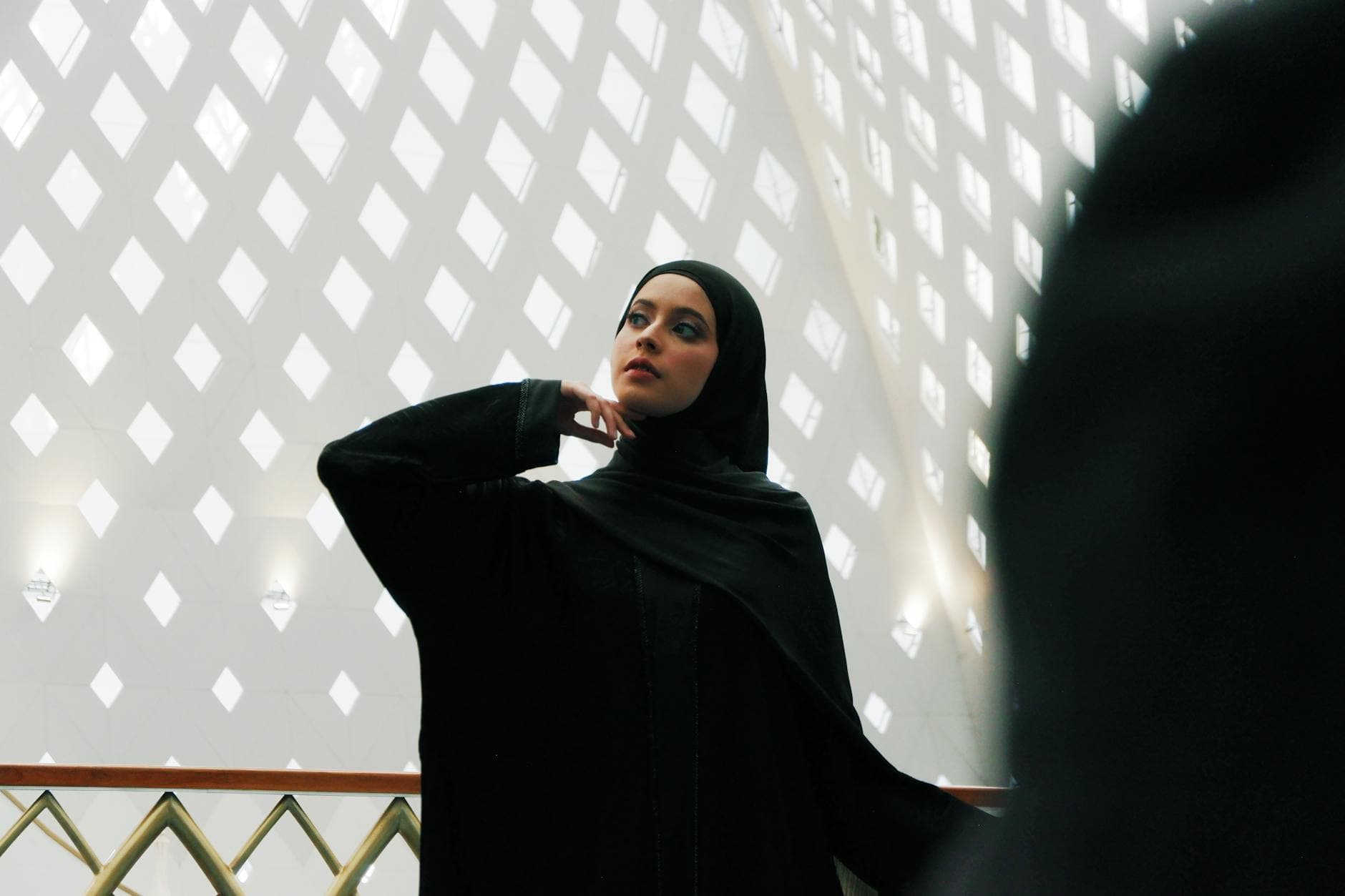 A thoughtful woman in hijab stands indoors against a patterned wall, capturing both elegance and cultural identity.