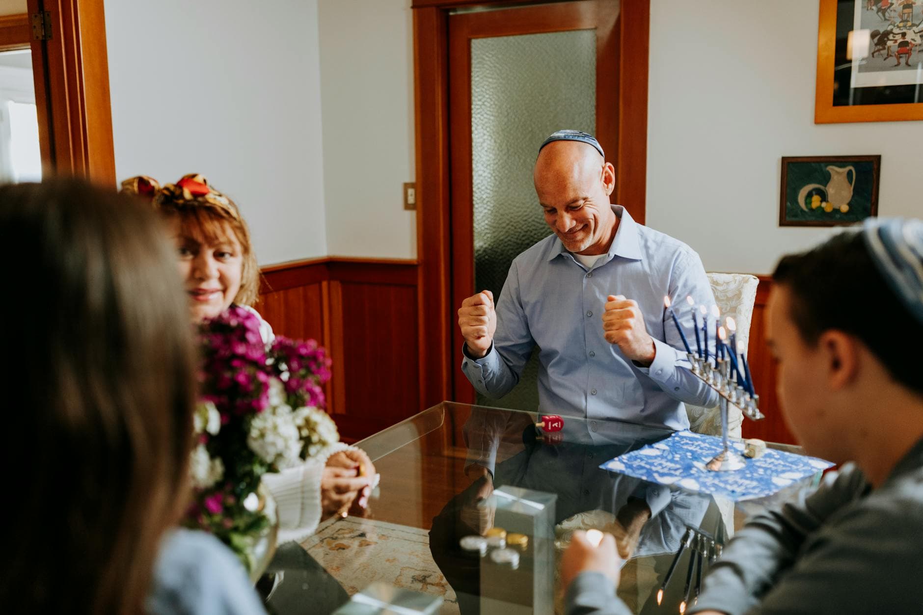 A family gathers around a glass table for a lively Hanukkah celebration with a menorah and dreidel.