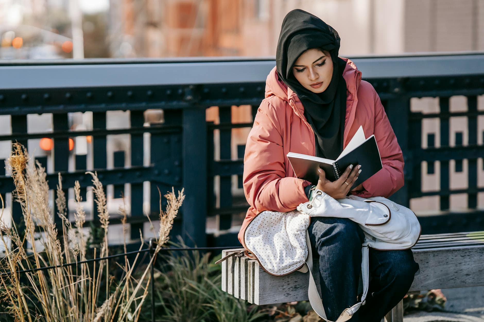 Muslim serious ethnic female in hijab and jacket analyzing information in notebook on street