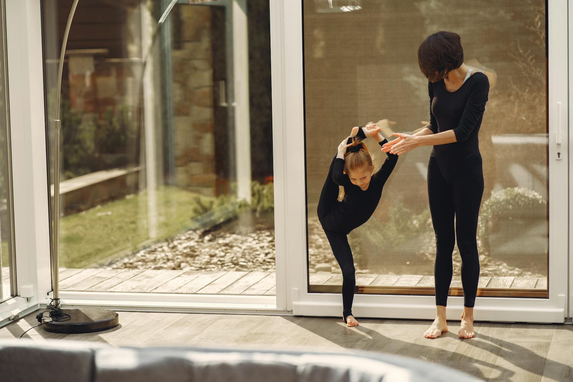 A mother helps her daughter practice ballet poses indoors by a sunny window.