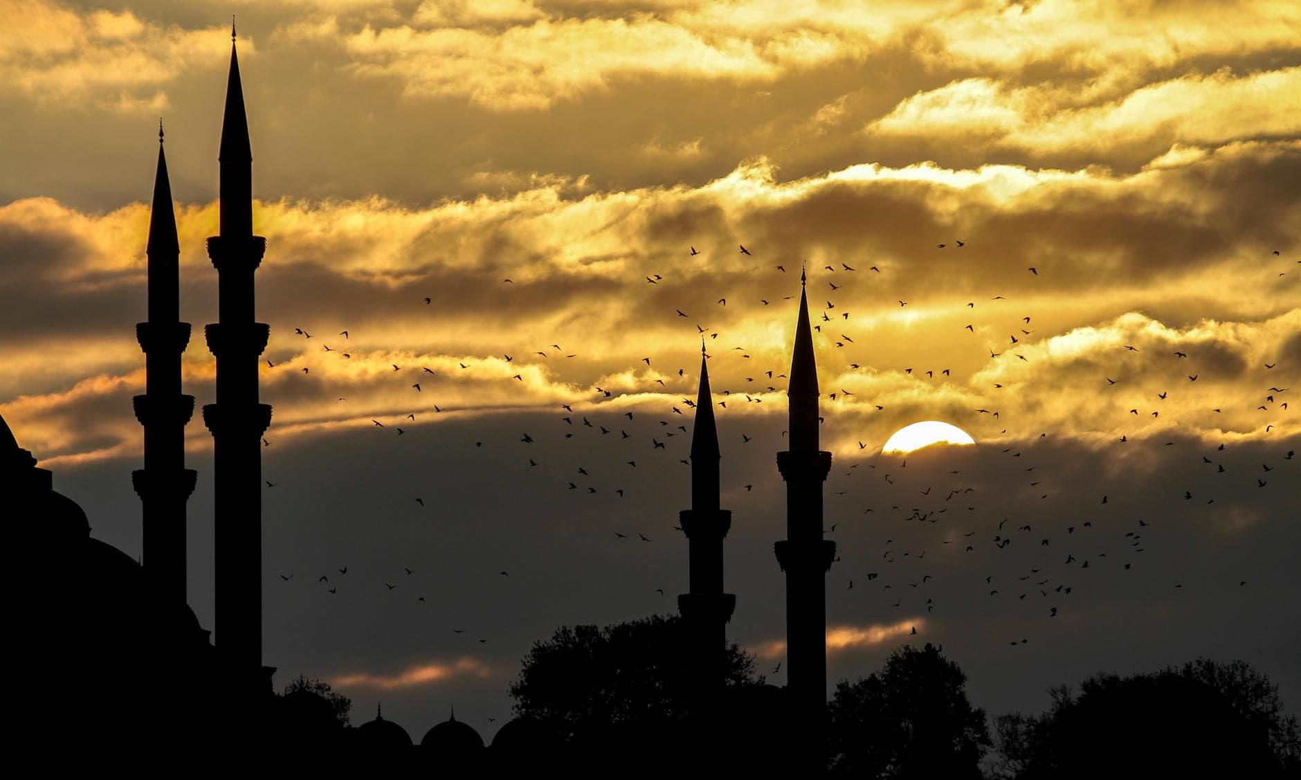 Silhouette of grand mosque towers against a dramatic sunset sky with birds in motion.