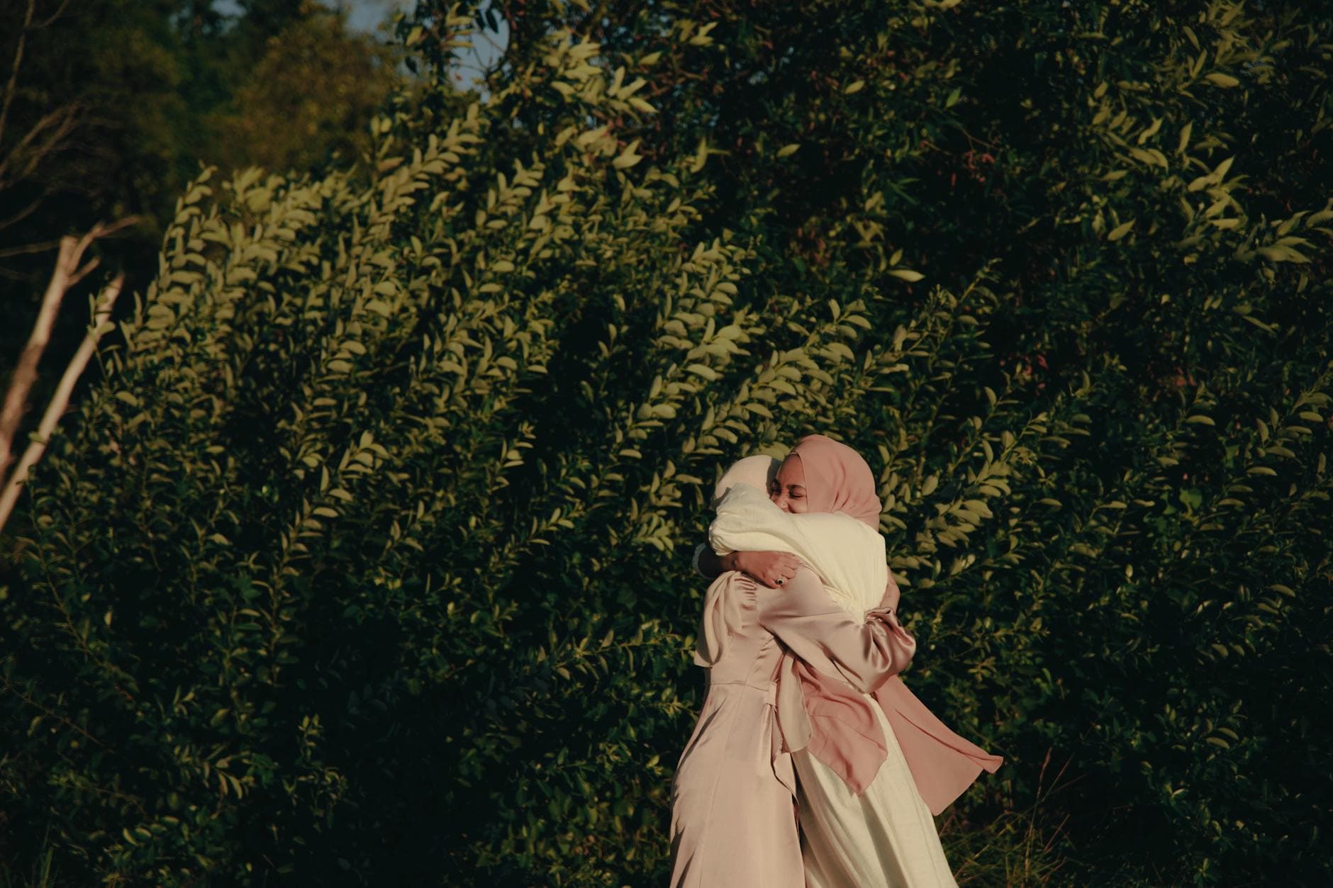 Two women in elegant dresses and hijabs share a heartfelt hug in a serene outdoor setting.