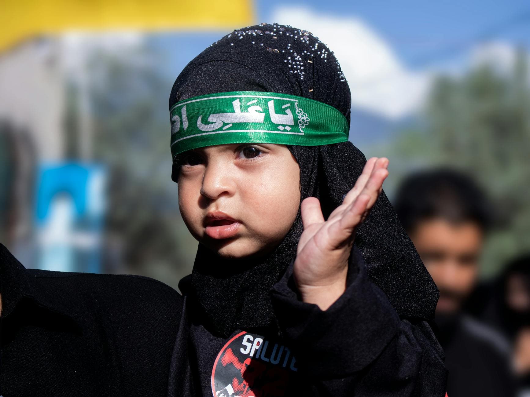 Child wearing a hijab with green headband during outdoor cultural event