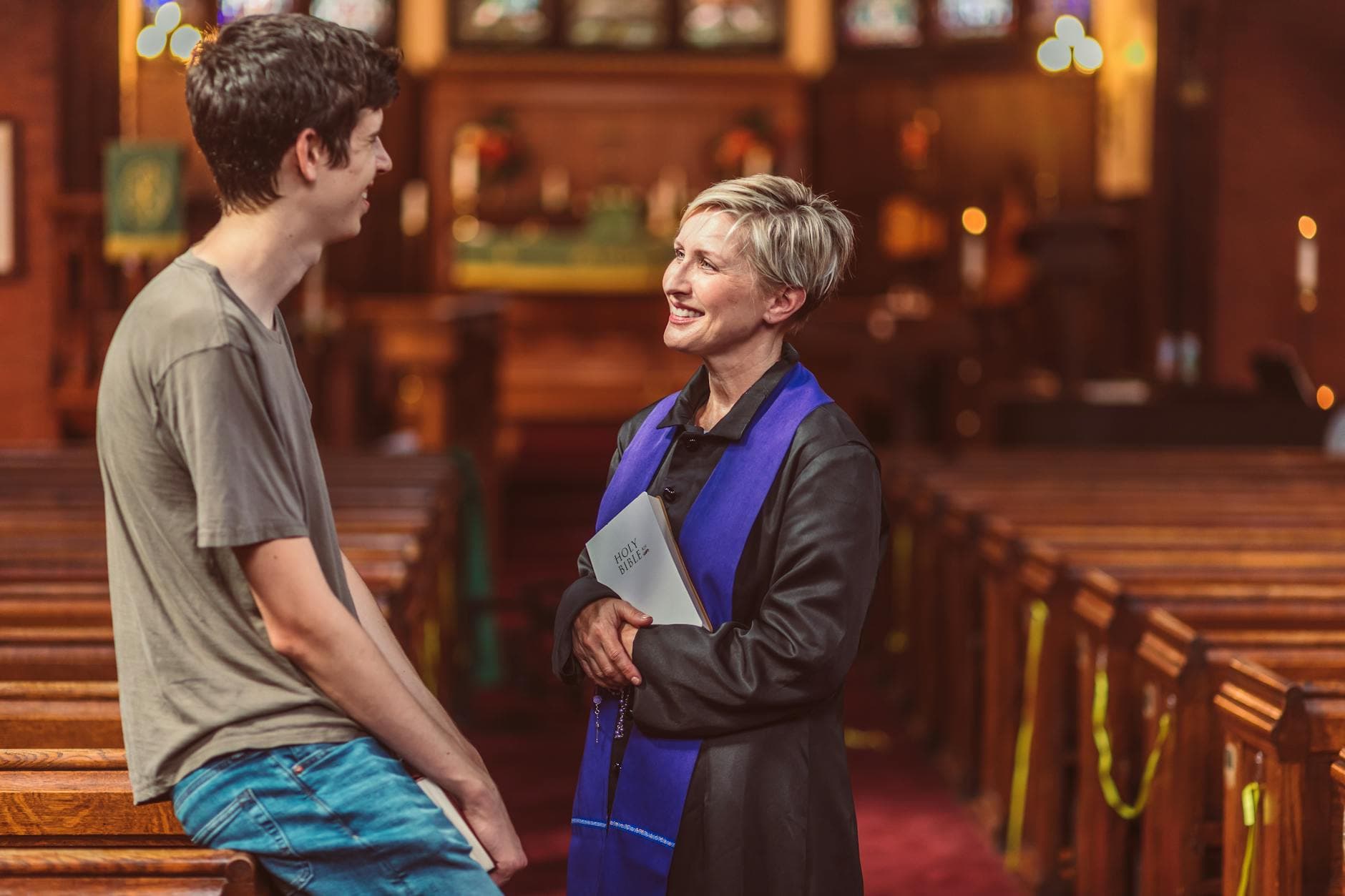 A priest holds a Bible while conversing with a young man in a warm, light-filled church interior.