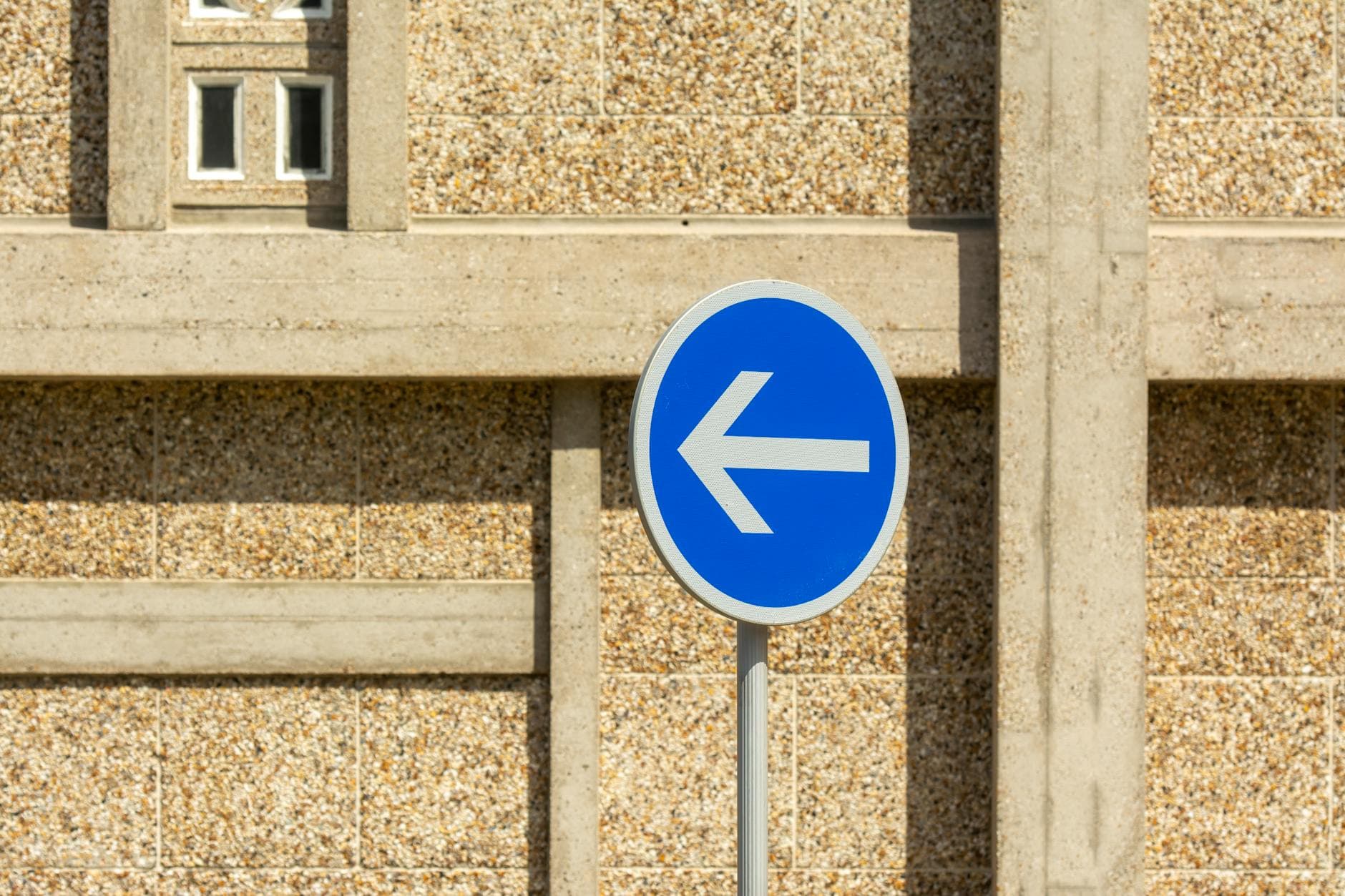 A blue traffic sign indicating a mandatory left turn in front of a textured urban wall.
